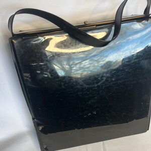 May include: A vintage black handbag with a glossy finish. The bag features a rectangular shape with a structured design, a top handle, and a clasp closure. The bag's surface reflects light, highlighting its sleek appearance. The handle is a curved, black strap.