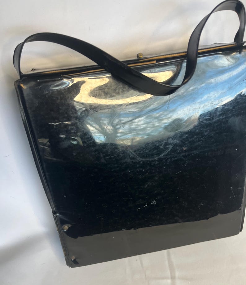 May include: A vintage black handbag with a glossy finish. The bag features a rectangular shape with a structured design, a top handle, and a clasp closure. The bag's surface reflects light, highlighting its sleek appearance. The handle is a curved, black strap.