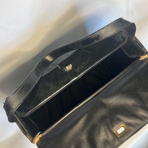 May include: Open black leather handbag with a top handle and a rectangular gold-toned clasp. The interior is black, and the bag appears to be vintage. The bag is open, revealing the inside.
