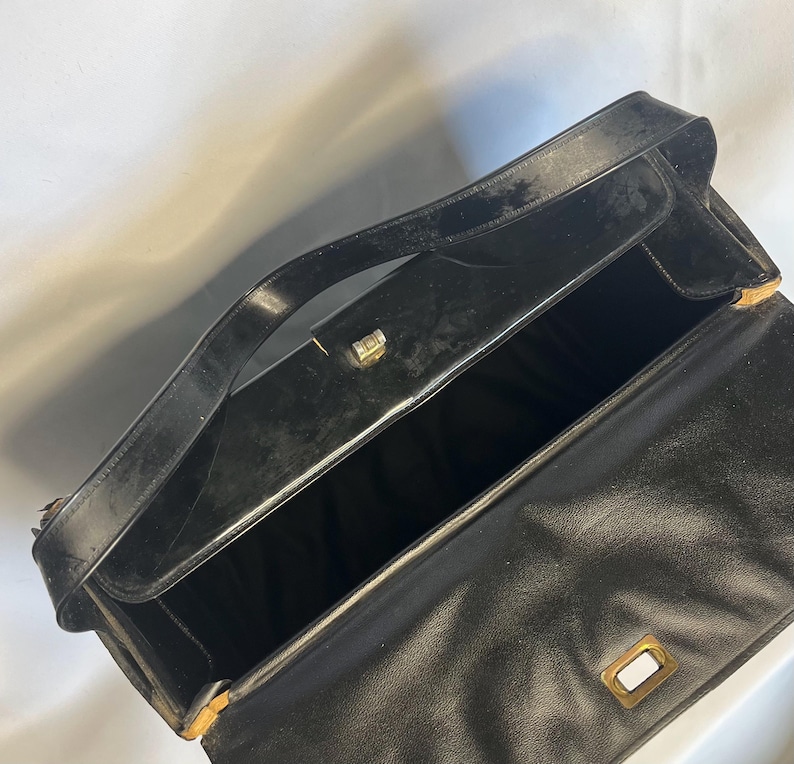 May include: Open black leather handbag with a top handle and a rectangular gold-toned clasp. The interior is black, and the bag appears to be vintage. The bag is open, revealing the inside.