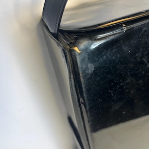 May include: A glossy black handbag with a rectangular shape and a single, curved handle. The bag's surface reflects light, and the handle appears to be made of a smooth material. The bag is set against a white background.