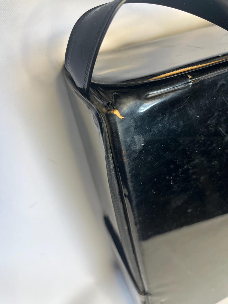 May include: A glossy black handbag with a rectangular shape and a single, curved handle. The bag's surface reflects light, and the handle appears to be made of a smooth material. The bag is set against a white background.