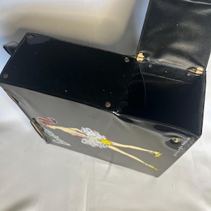 May include: Black vintage handbag with a glossy finish. The bag features a cartoon illustration of a woman in a dress and yellow shoes. The bag has a handle and a hinged lid that opens to reveal a black interior.