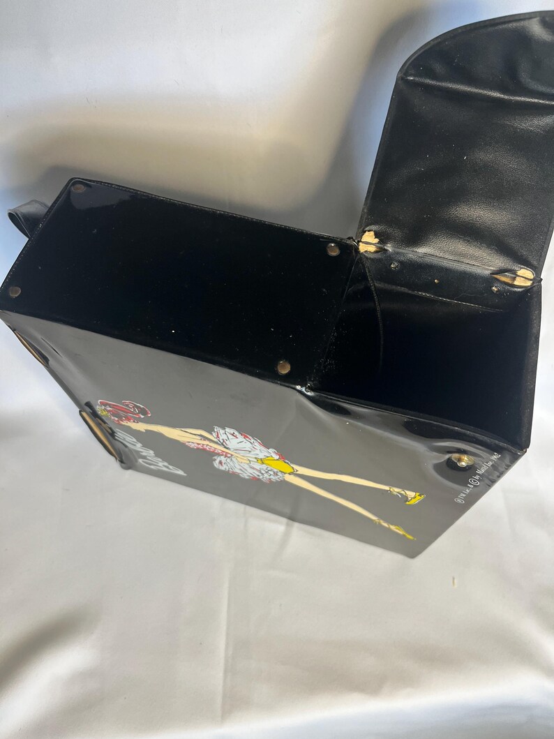 May include: Black vintage handbag with a glossy finish. The bag features a cartoon illustration of a woman in a dress and yellow shoes. The bag has a handle and a hinged lid that opens to reveal a black interior.