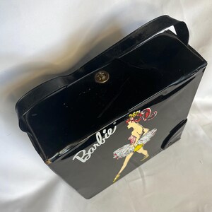 May include: Black vintage Barbie carrying case with a handle and a gold-colored clasp. The case features a graphic of Barbie in a ballerina outfit with the word "Barbie" in white script.