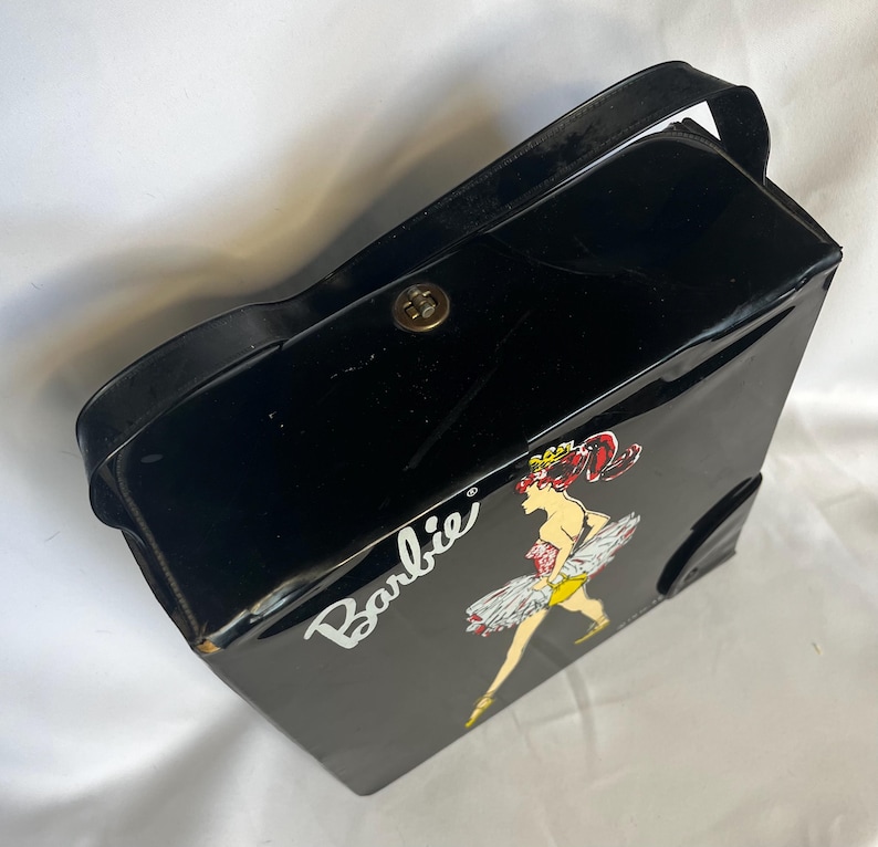 May include: Black vintage Barbie carrying case with a handle and a gold-colored clasp. The case features a graphic of Barbie in a ballerina outfit with the word "Barbie" in white script.