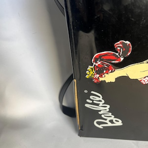 May include: Black Barbie-branded box with a cartoon graphic of a woman in a red outfit. The word "Barbie" is written in white cursive. A black strap is attached to the box.