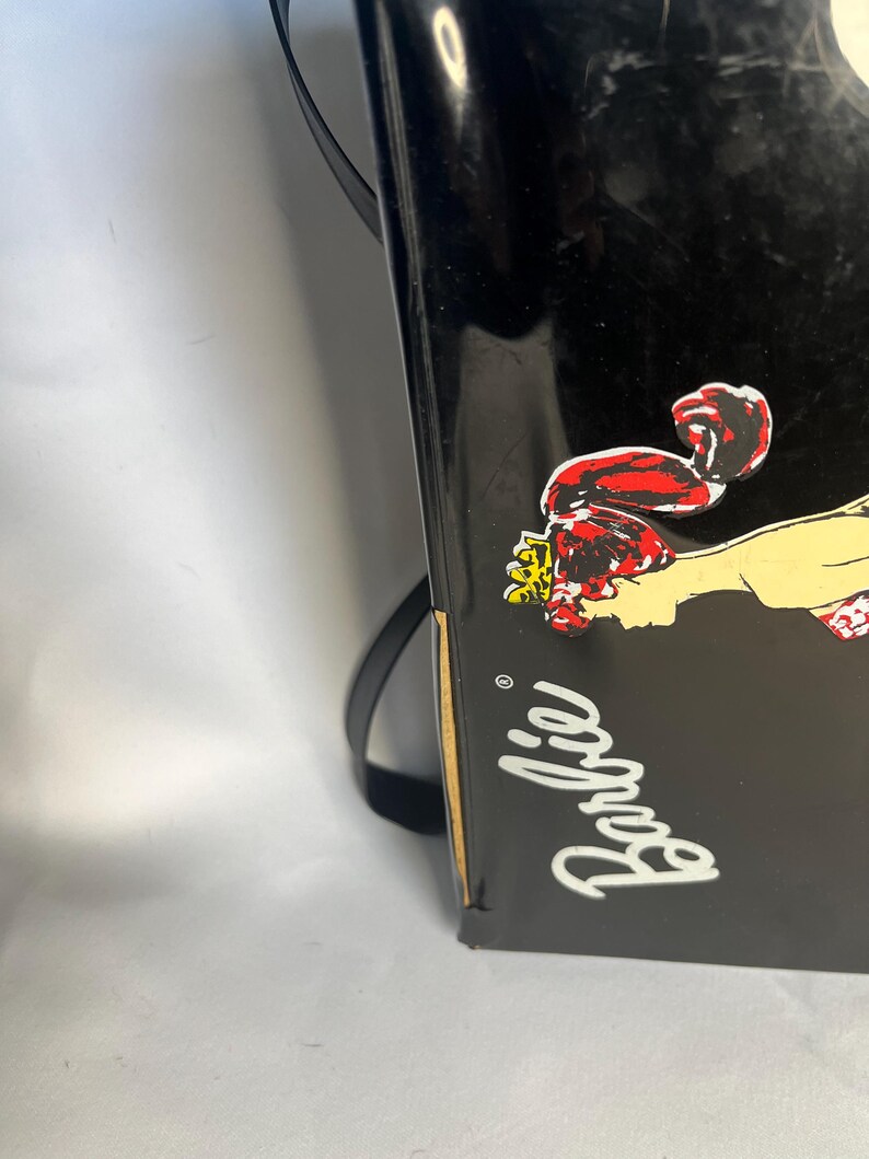 May include: Black Barbie-branded box with a cartoon graphic of a woman in a red outfit. The word "Barbie" is written in white cursive. A black strap is attached to the box.
