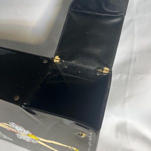May include: Black vintage box with a hinged lid and a clasp. The box has a glossy finish and a decorative illustration of a woman in a dress. The lid is open, revealing the interior. The box is likely made of a hard material.
