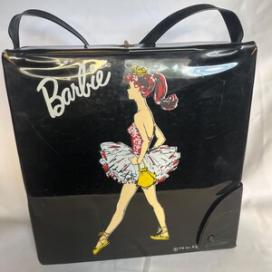 May include: Black vintage Barbie tote bag with a ballerina graphic. The bag features a black body with a glossy finish, black handles, and a snap closure. The Barbie logo is in white.