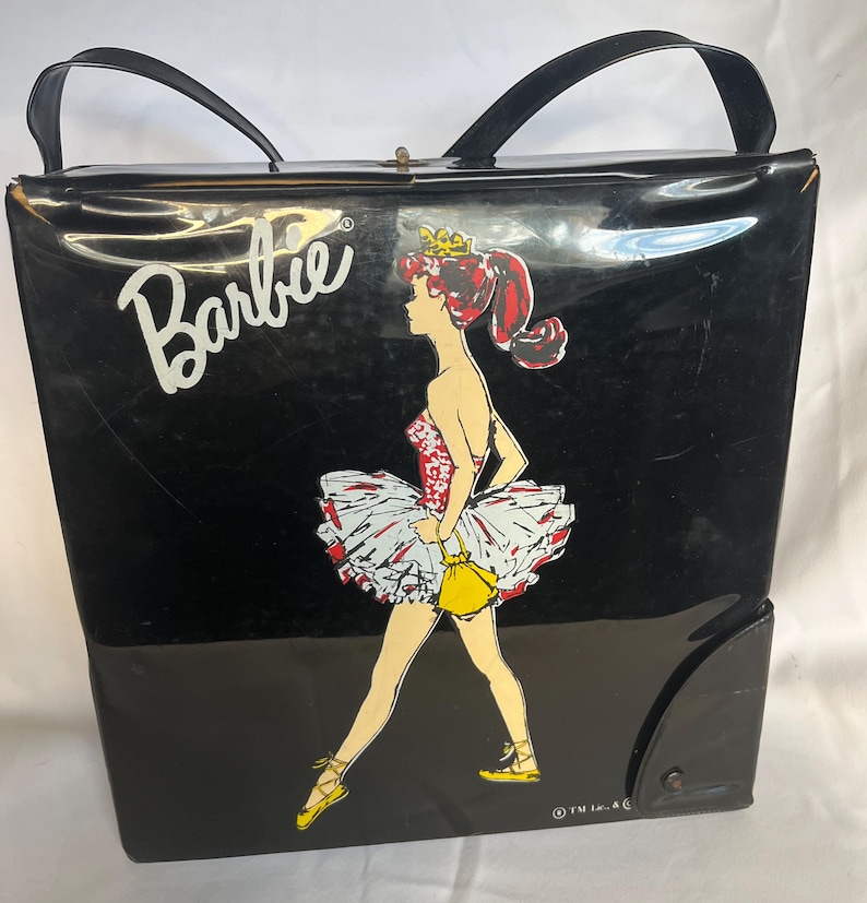 May include: Black vintage Barbie tote bag with a ballerina graphic. The bag features a black body with a glossy finish, black handles, and a snap closure. The Barbie logo is in white.