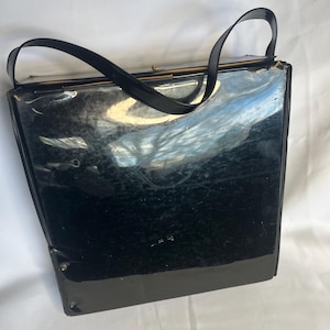 May include: A vintage black handbag with a glossy finish and two black handles. The rectangular purse has a clasp closure and appears to be made of a patent leather or similar material. The bag shows signs of wear.