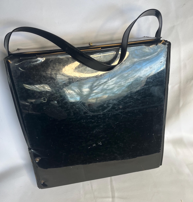 May include: A vintage black handbag with a glossy finish and two black handles. The rectangular purse has a clasp closure and appears to be made of a patent leather or similar material. The bag shows signs of wear.