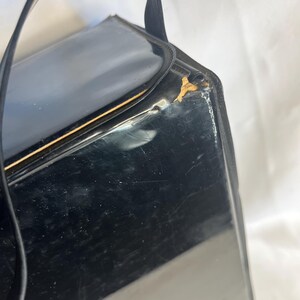 May include: A vintage black handbag with a glossy finish and a curved handle. The rectangular purse has a gold-colored trim and shows signs of wear. The handle is made of a matching black material.