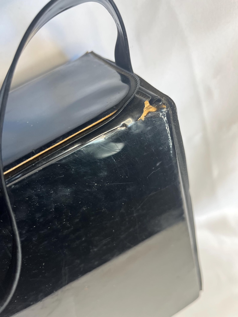 May include: A vintage black handbag with a glossy finish and a curved handle. The rectangular purse has a gold-colored trim and shows signs of wear. The handle is made of a matching black material.