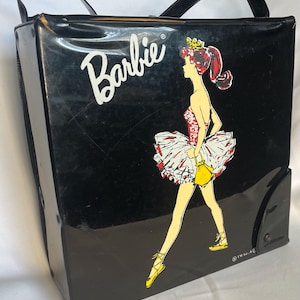 May include: Black vintage Barbie carrying case with a ballerina graphic. The case features a white "Barbie" logo and a ballerina in a red, white, and yellow outfit. It has a black handle and a rectangular shape.
