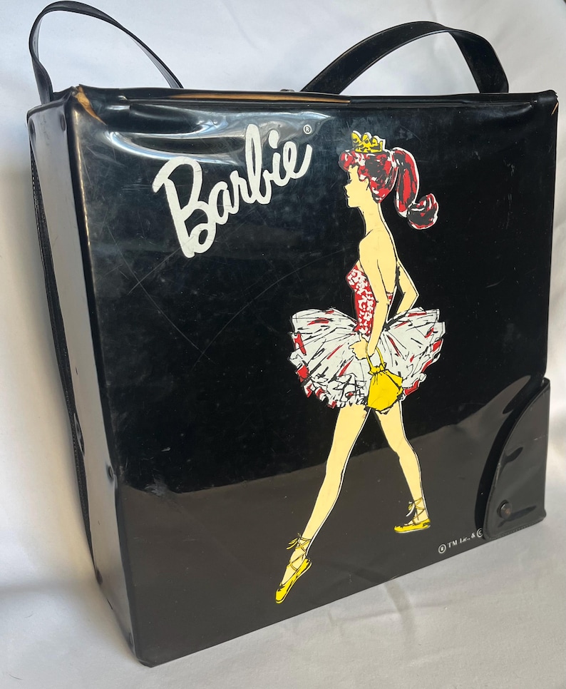 May include: Black vintage Barbie carrying case with a ballerina graphic. The case features a white "Barbie" logo and a ballerina in a red, white, and yellow outfit. It has a black handle and a rectangular shape.