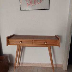 Puede incluir: Una consola de madera de estilo moderno de mediados de siglo con dos cajones y pomos de latón. La mesa tiene patas cónicas y una parte superior rectangular. La obra de arte sobre la mesa tiene el texto "RON MUECK".