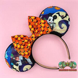 May include: Halloween-themed mouse ears headband with a large orange bow featuring black bat silhouettes. The ears display Mickey Mouse and other characters against a spooky night sky backdrop. The headband is black with a brown interior.