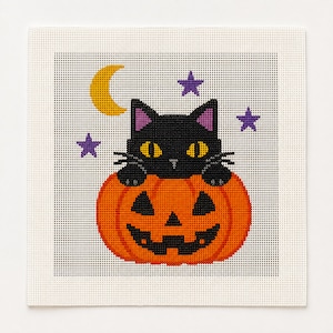 May include: A square cross-stitch pattern on white fabric featuring a black cat with yellow eyes peeking out from behind an orange jack-o'-lantern. The design includes a yellow crescent moon and three purple stars arranged in the background.