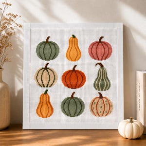 May include: A square cross-stitch embroidery piece featuring a grid of nine pumpkins in various shapes and colors, including green, orange, yellow, and cream, set against a white Aida cloth background and framed in a simple white wooden frame.