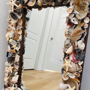 May include: A rectangular mirror with a dark wooden frame completely covered in a dense arrangement of various seashells and small white starfish. The shells display a natural palette of beige, brown, grey, and white tones in diverse shapes.