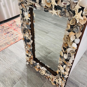 May include: A rectangular mirror featuring a dark brown frame densely covered in a variety of natural seashells and small white starfish. The shells vary in size, shape, and color, including shades of white, beige, tan, and grey, creating a textured border around the reflective glass surface.