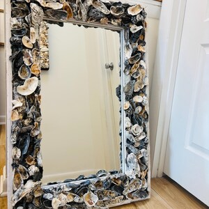 Puede incluir: Un espejo rectangular con marco blanco, decorado con conchas de ostra en varios tonos. Las conchas, en blanco, gris y marrón, crean un borde texturizado alrededor del cristal reflectante. El espejo está apoyado contra una pared.