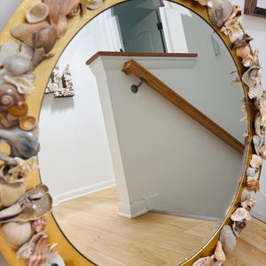 May include: A circular mirror with a gold frame, decorated with assorted seashells. The mirror reflects a staircase and wooden floor. The seashells are in shades of white, beige, and brown.