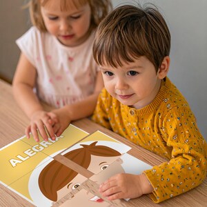 May include: Two young children are focused on assembling a puzzle. The puzzle pieces form a cartoon face with the word "ALEGRIA" visible. The puzzle is on a wooden surface. One child wears a yellow sweater, and the other wears a white shirt.