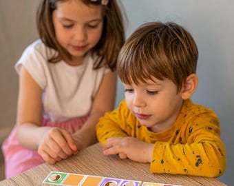 Puede incluir: Dos niños jugando a un juego de cartas sobre una mesa de madera. El juego presenta cartas coloridas con ilustraciones de caras y texto. Los niños están concentrados en el juego, con las cartas dispuestas frente a ellos.
