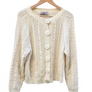 May include: A cream-coloured long-sleeve cardigan sweater featuring a chunky cable knit pattern, a round neckline, and five large, round, matching cream buttons down the front. The garment is displayed on a wooden hanger against a white background.