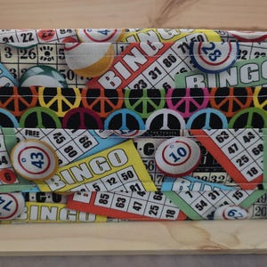 May include: A rectangular fabric storage box with a colorful bingo-themed pattern. The design includes bingo cards, bingo balls, and the word "BINGO" in various fonts and colors. A red polka-dot fabric tab is attached to the side.