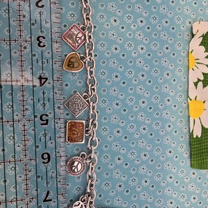 May include: Silver charm bracelet with various charms, including a peace sign, a heart, and a square with a design. The bracelet is approximately 8 inches long. The charms have colorful accents and the bracelet is on a blue floral background.