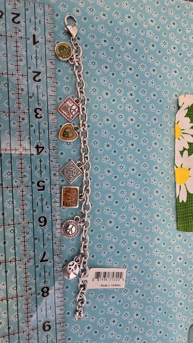 May include: Silver charm bracelet with various charms, including a peace sign, a heart, and a square with a design. The bracelet is approximately 8 inches long. The charms have colorful accents and the bracelet is on a blue floral background.