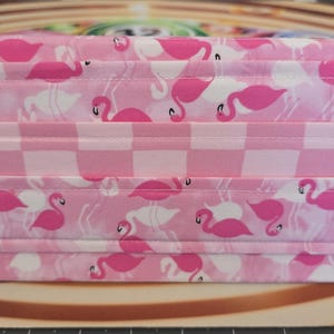May include: A pink fabric container with a flamingo pattern. The design features pink flamingos on a white background, interspersed with a pink and white checkered pattern. The container is rectangular and appears to be folded.