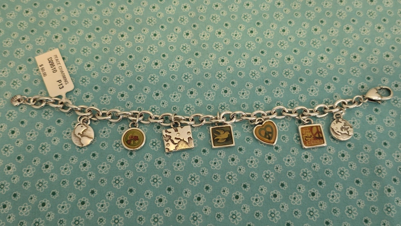 May include: Silver charm bracelet with various charms, including a dove, a peace sign, and a world map. The bracelet has a lobster clasp closure and a price tag that reads "PEACE CHARMS". The charms are in various shapes and colors.