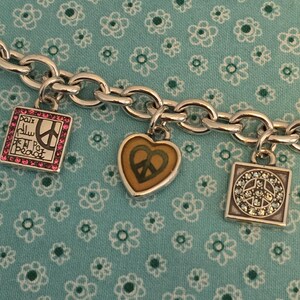 May include: Silver-toned charm bracelet with three charms. One charm is a square with pink gems and the word "Peace" in multiple languages. Another is a heart with a peace sign. The third is a square with a peace sign and clear gems.