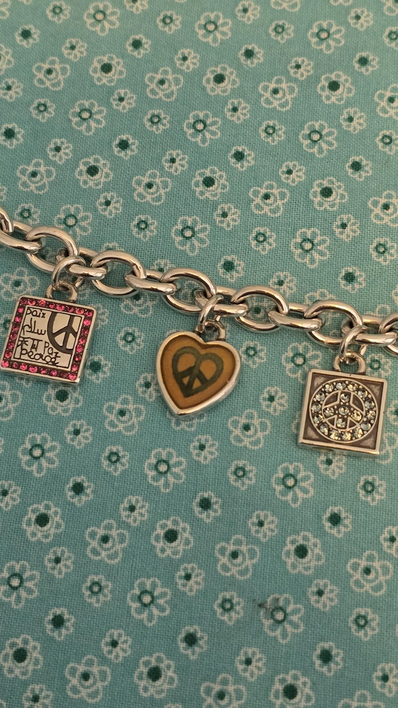May include: Silver-toned charm bracelet with three charms. One charm is a square with pink gems and the word "Peace" in multiple languages. Another is a heart with a peace sign. The third is a square with a peace sign and clear gems.