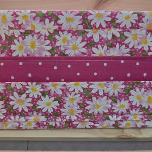 May include: A rectangular fabric storage bin with a pink daisy floral pattern. A solid pink band with white polka dots accents the center. The bin is made of quilted fabric and sits on a wooden surface.