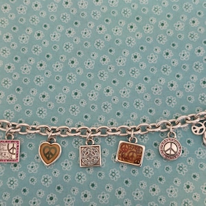 May include: Silver charm bracelet featuring various peace-themed charms. Charms include a peace sign, heart, and square designs. The bracelet is displayed on a light blue background with a floral pattern.