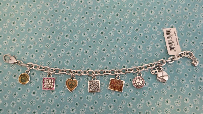 May include: Silver charm bracelet featuring various peace-themed charms. Charms include a peace sign, heart, and square designs. The bracelet is displayed on a light blue background with a floral pattern.