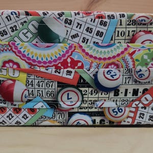 Op de afbeelding: Een stoffen opbergdoos met een kleurrijk bingomotief. De doos heeft bingoballen en -kaarten, met een paarse stoffen handgreep. Het ontwerp bevat nummers, patronen en het woord "BINGO".