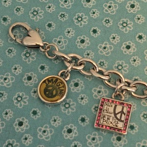 May include: Silver-toned charm bracelet with three charms. One charm reads "IMAGINE" with a peace sign, another has the word "Peace" in multiple languages, and the third is a peace sign. The bracelet is on a blue floral background.