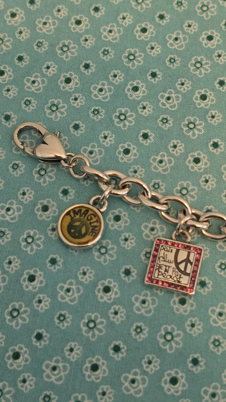 May include: Silver-toned charm bracelet with three charms. One charm reads "IMAGINE" with a peace sign, another has the word "Peace" in multiple languages, and the third is a peace sign. The bracelet is on a blue floral background.