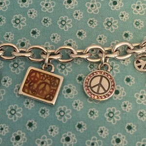 May include: Silver charm bracelet with multiple charms. Charms include a square with a peace sign, a round charm with a peace sign surrounded by pink stones, and a peace sign charm. The bracelet is on a blue floral background.