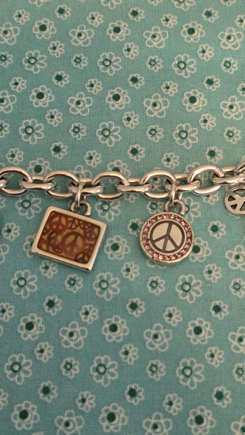 May include: Silver charm bracelet with multiple charms. Charms include a square with a peace sign, a round charm with a peace sign surrounded by pink stones, and a peace sign charm. The bracelet is on a blue floral background.