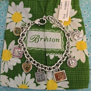May include: A silver charm bracelet with various charms, including peace signs, hearts, and squares. The bracelet is displayed on a green daisy-patterned bag with the brand name "Brighton" visible. The bag is on a light blue floral background.