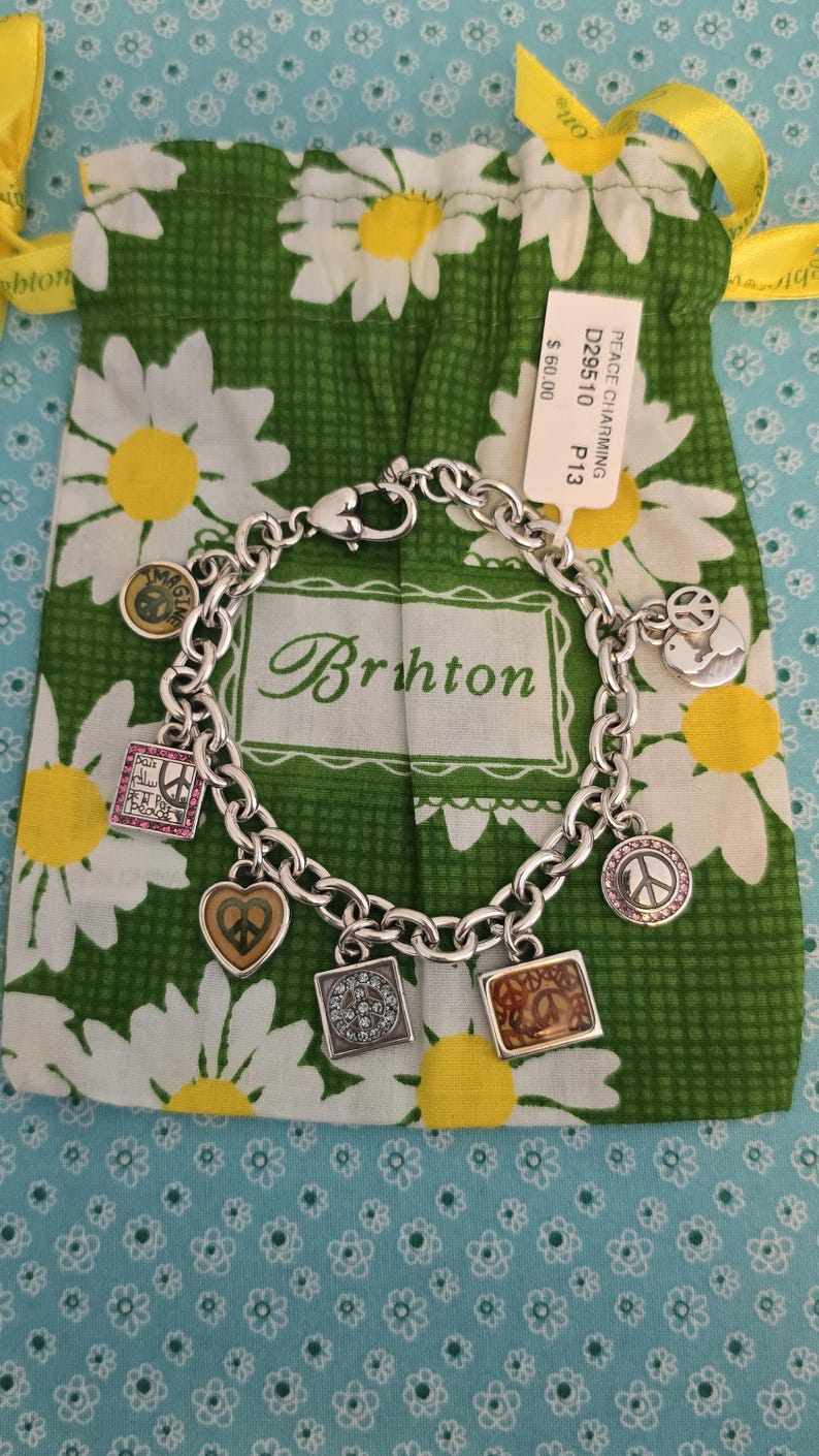 May include: A silver charm bracelet with various charms, including peace signs, hearts, and squares. The bracelet is displayed on a green daisy-patterned bag with the brand name "Brighton" visible. The bag is on a light blue floral background.