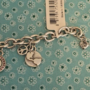 May include: Silver-toned charm bracelet with multiple charms, including a peace sign, a globe, and a heart-shaped charm. The bracelet is displayed on a teal fabric with a floral pattern. A tag with a barcode is visible.